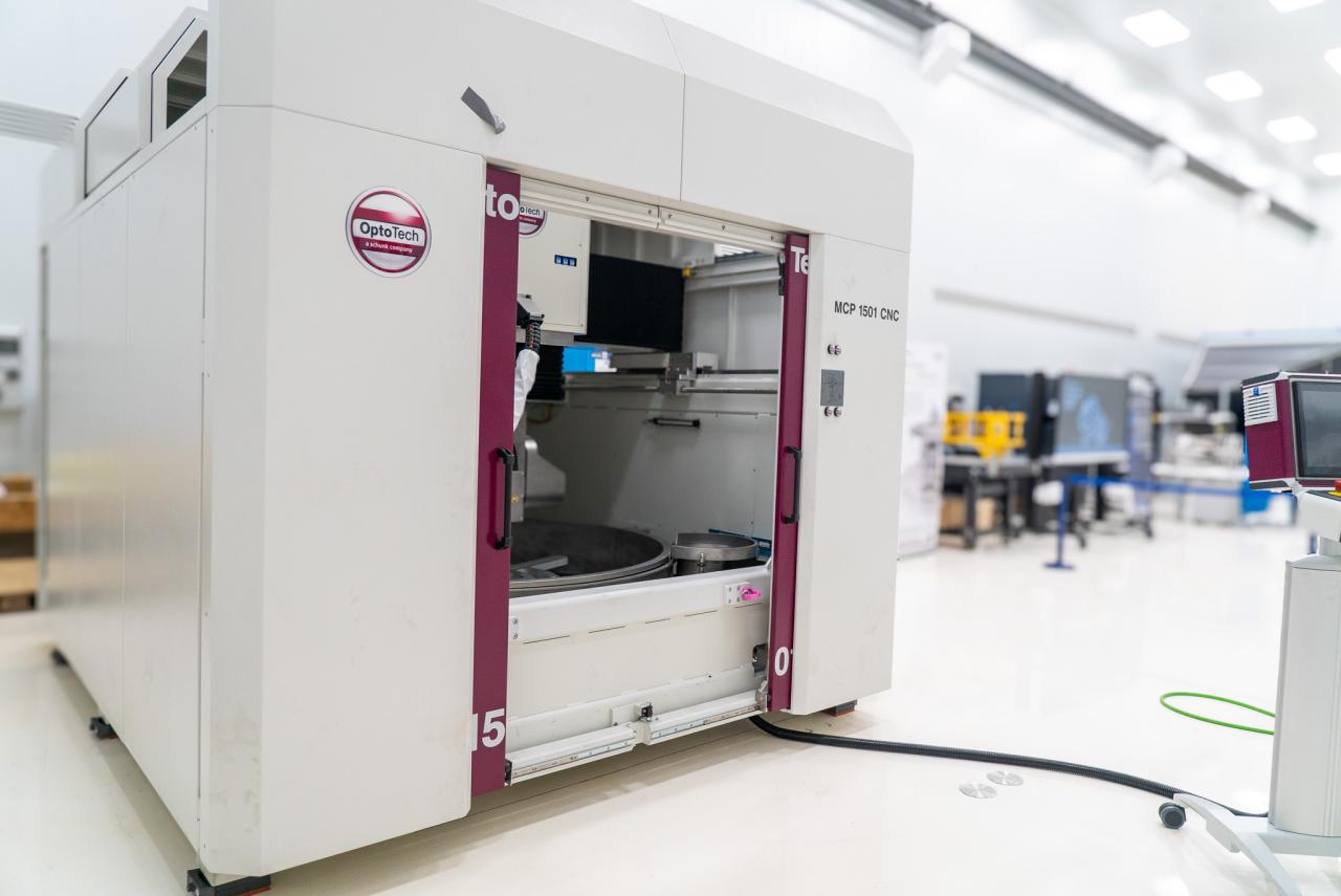 A large white machine with two big doors (polishing machine for large surfaces) in a white room with high ceilings (IACTEC's south clean room).