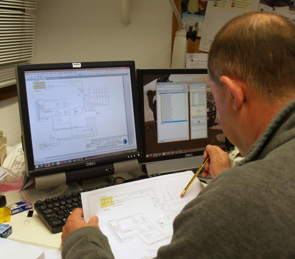 Vista de un técnico trabajando en un diseño electrónico con un plano y la pantalla del ordenador