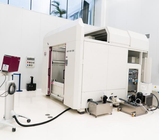 A large white machine with two big doors (polishing machine for large surfaces) in a white room with high ceilings (IACTEC's south clean room).