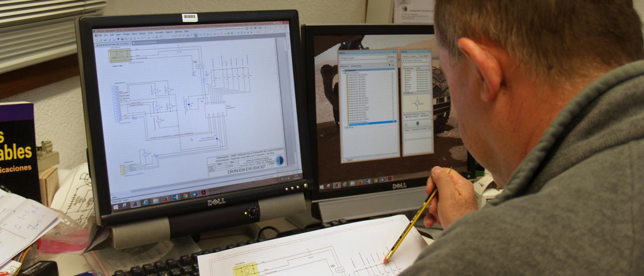 Vista de un técnico trabajando en un diseño electrónico con un plano y la pantalla del ordenador