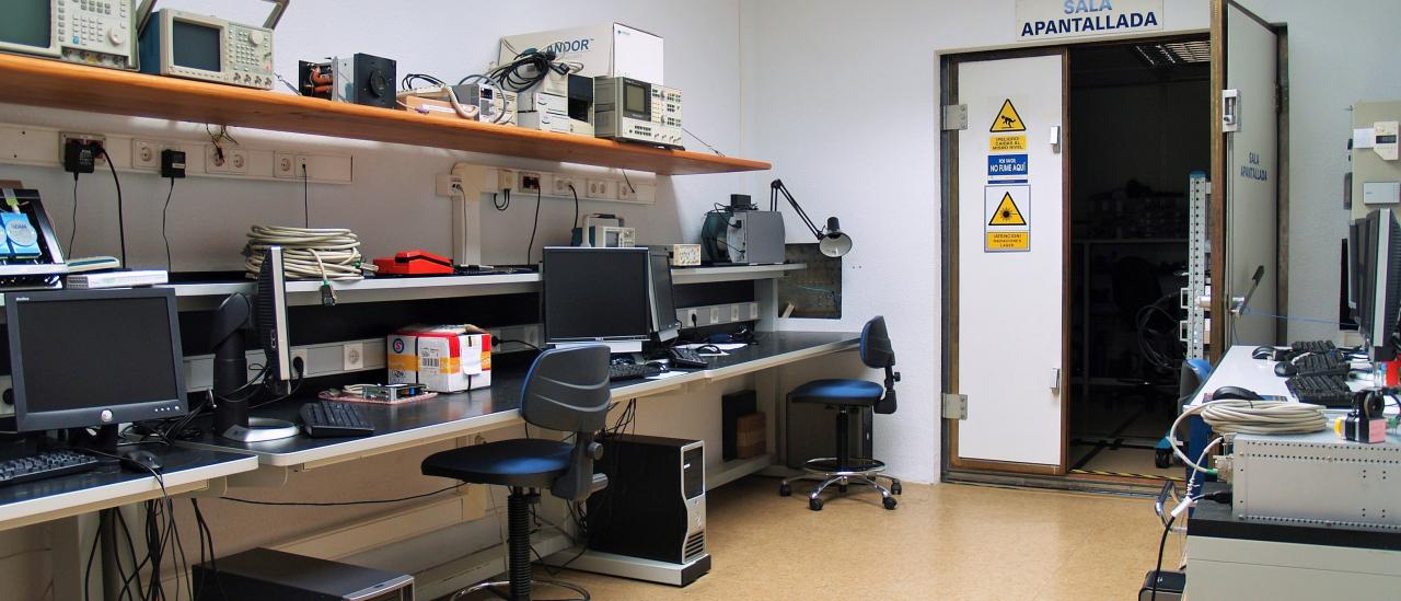 Vista general del laboratorio de compatibilidad electromagnética con bancos de trabajo y ordenadores y vista al fondo la sala de la puerta de entrada a la sala apantallada