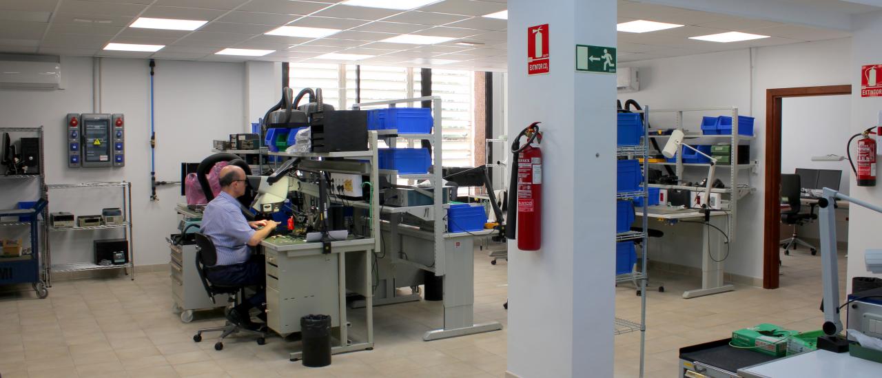 Vista general del Taller de Electrónica con varios bancos de trabajo, armarios y cajas para almacenar componentes y piezas electrónicas, y un técnico trabajando en uno de los bancos.