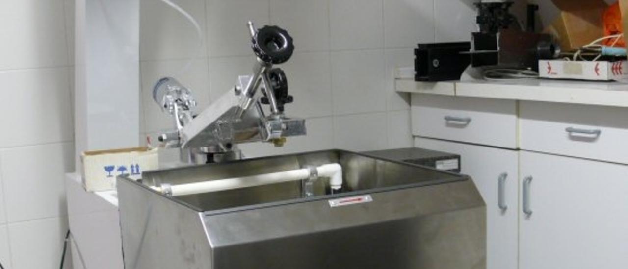 Imagen de una máquina de pulido óptico en el laboratorio. Máquina cuadrada con un seno a media altura para colocar los materiales a pulir y con unas abrazaderas metálicas por encima para sujetar la pieza pulidora. Los mandos para el control de máquina están en la parte alta.