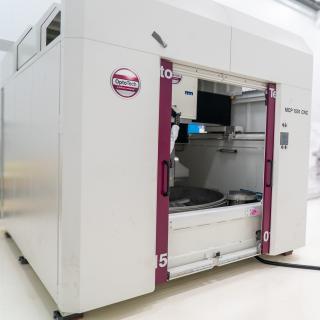 A large white machine with two big doors (polishing machine for large surfaces) in a white room with high ceilings (IACTEC's south clean room).