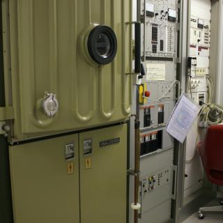 Vista de la evaporadora y de su electrónica asociada en el laboratorio. Máquina con una puerta reforzada y un ojo de buey, y dos grandes racks con componentes electrónicos, botones e indicadores para el control de la máquina.