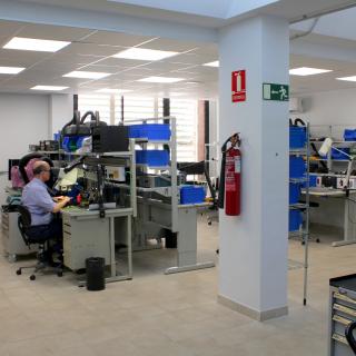 Vista general del Taller de Electrónica con varios bancos de trabajo, armarios y cajas para almacenar componentes y piezas electrónicas, y un técnico trabajando en uno de los bancos.