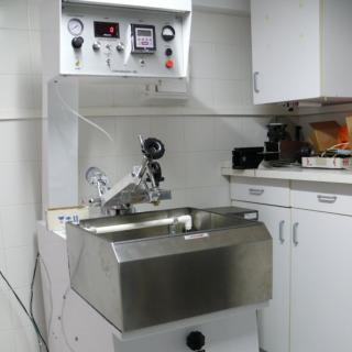 Imagen de una máquina de pulido óptico en el laboratorio. Máquina cuadrada con un seno a media altura para colocar los materiales a pulir y con unas abrazaderas metálicas por encima para sujetar la pieza pulidora. Los mandos para el control de máquina están en la parte alta.